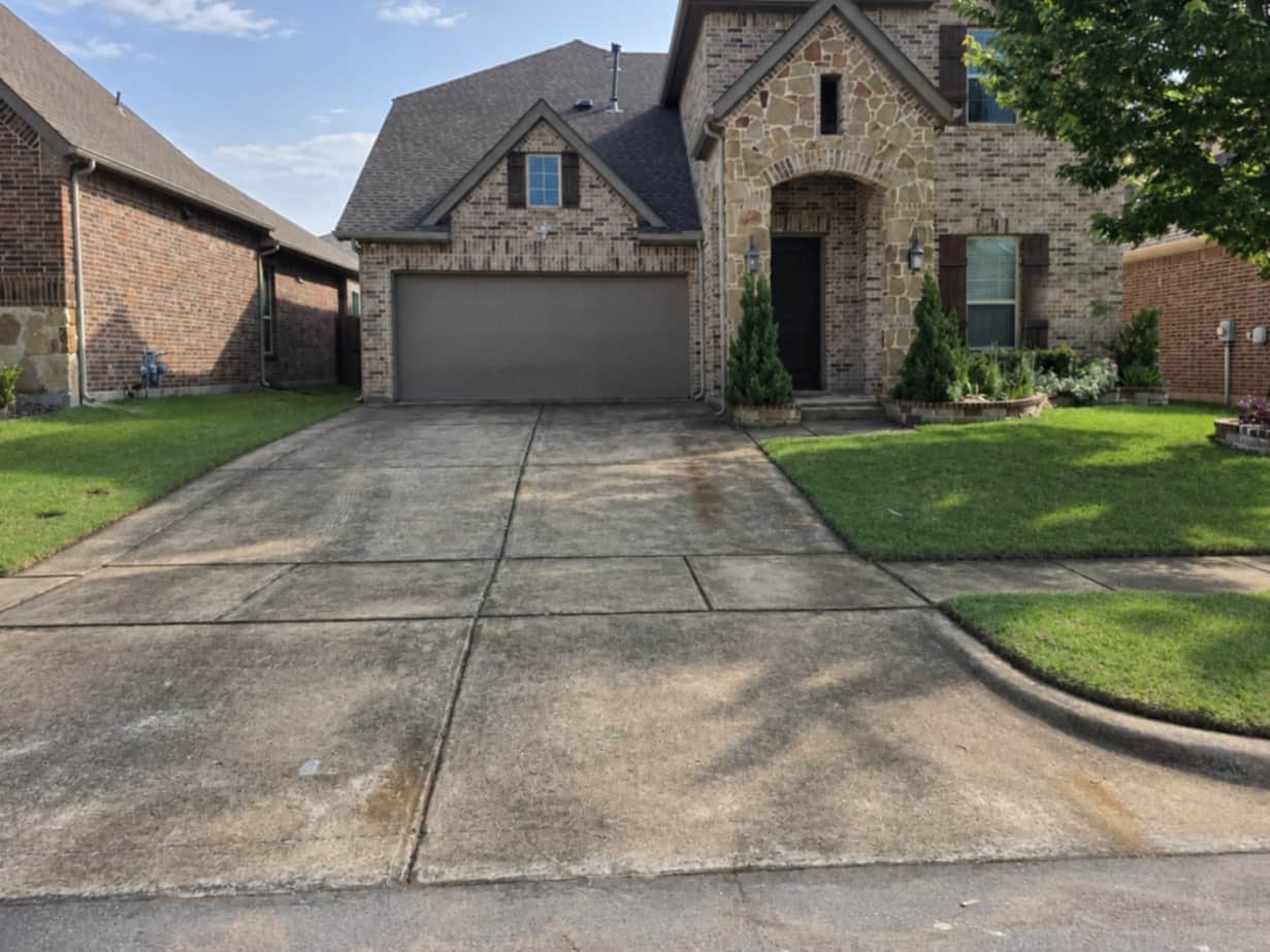 Concrete driveway of a two-story brick and stone home, darkened across its whole width with rust-coloured runoff, tire marks and organic staining before cleaning.